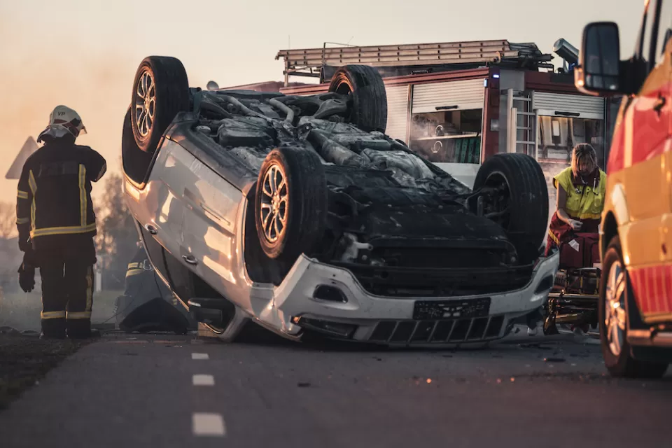 Car crash scene with overturned car, emergency vehicles, and firefighter standing at left.
